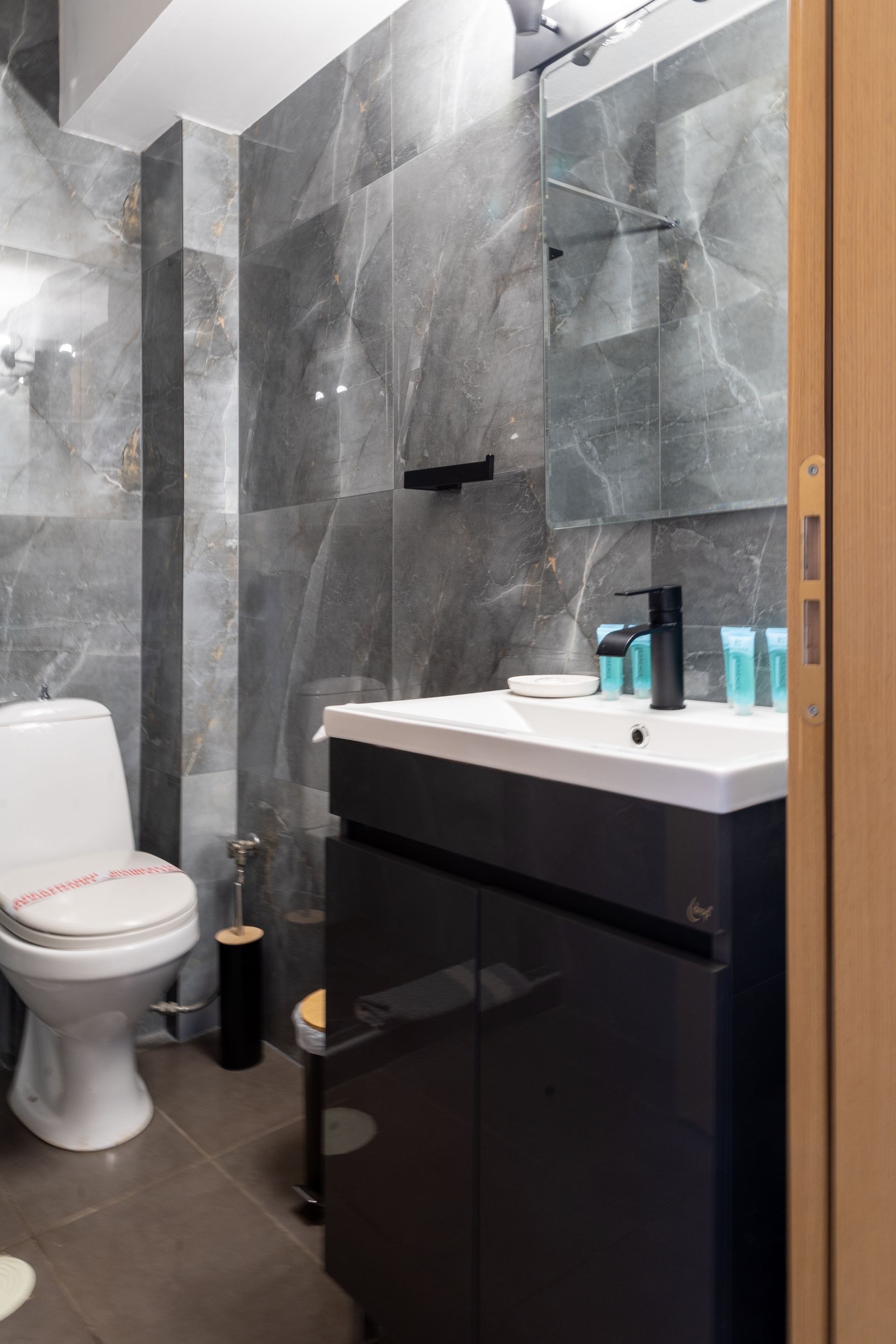 Compact modern bathroom with gray tile shower, white toilet, and dark vanity with sink and toiletries