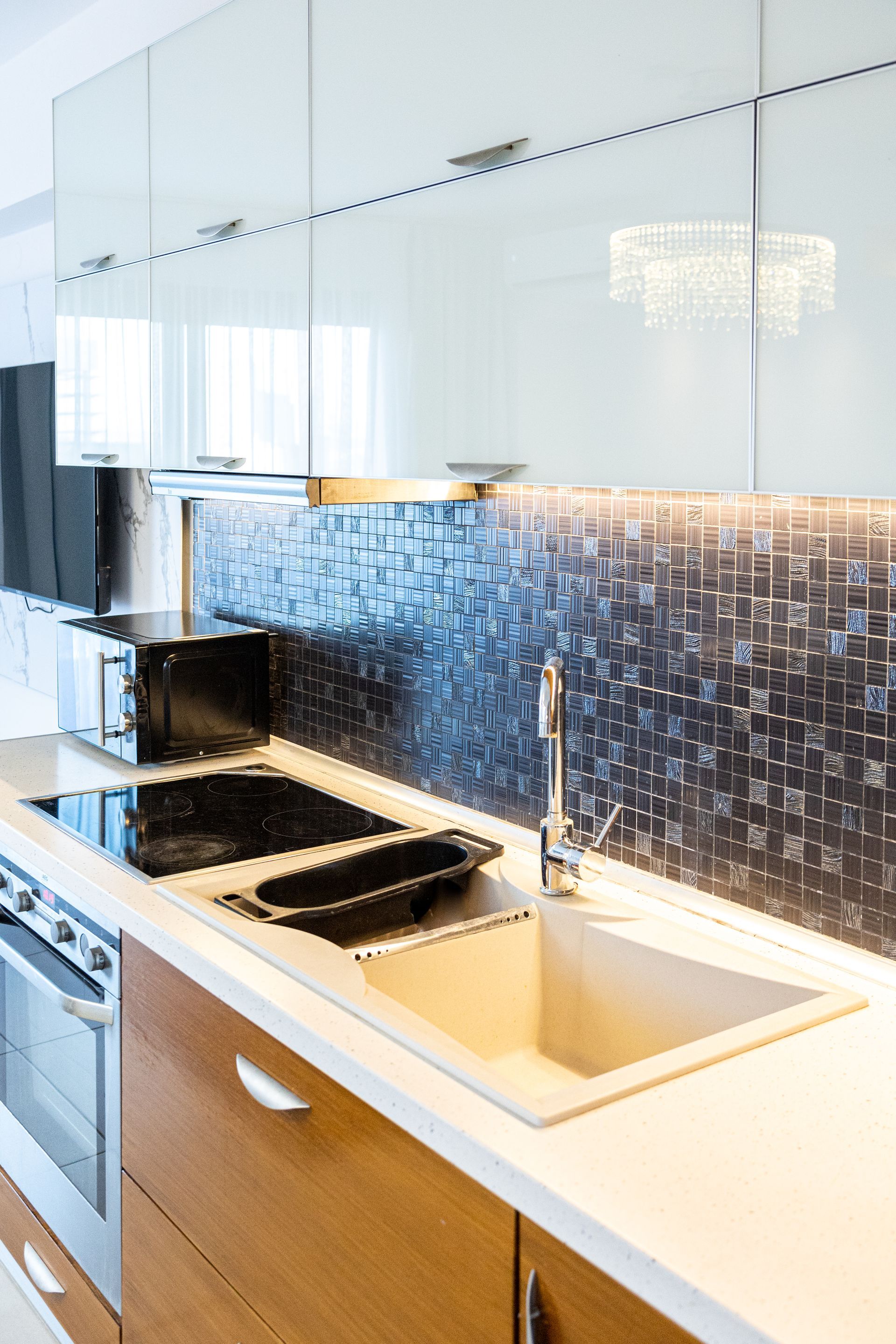 Modern kitchen sink with white countertop, wood cabinetry, and blue mosaic backsplash under glossy cabinets