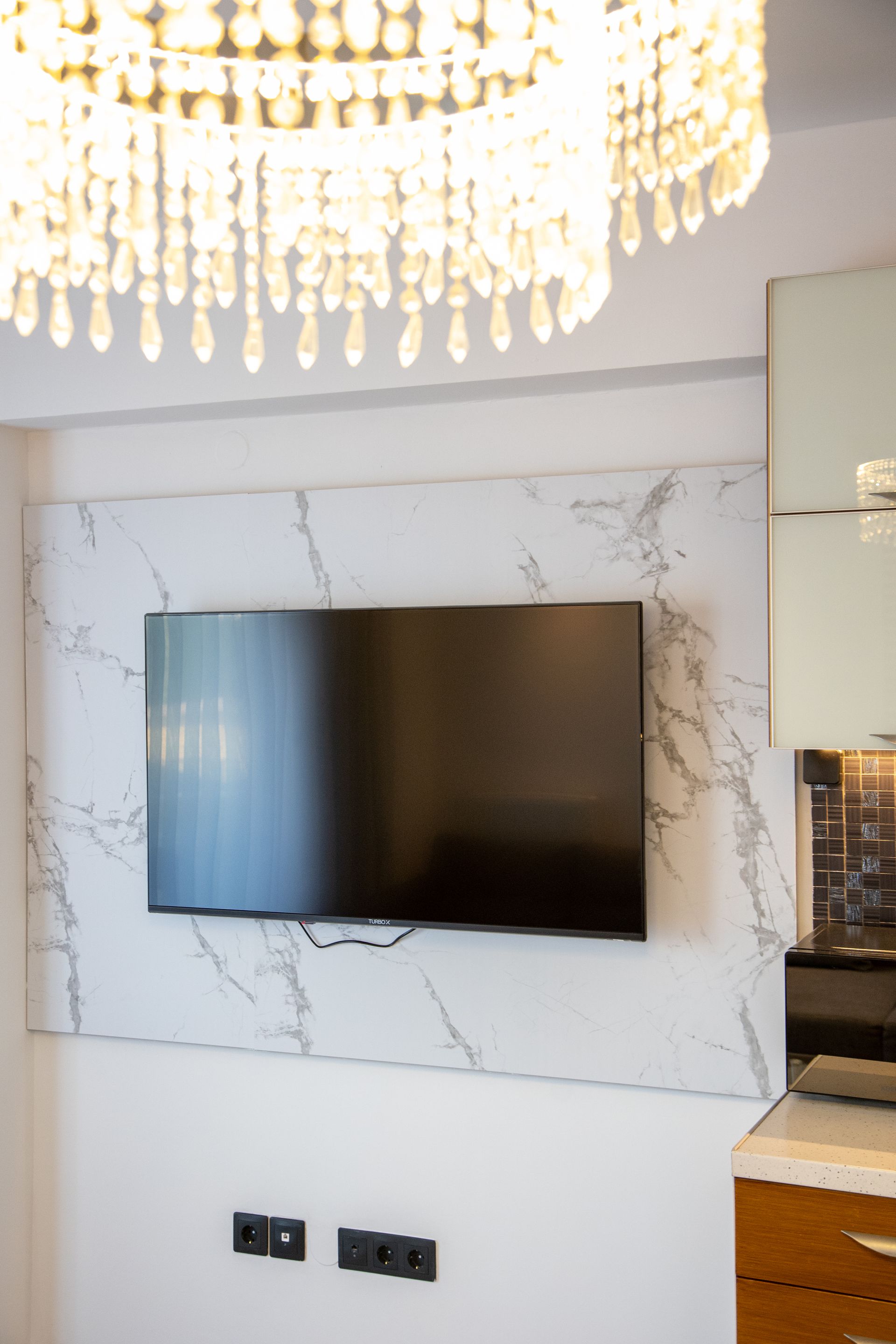 Wall-mounted TV on a marble accent wall beneath a crystal chandelier in a modern room