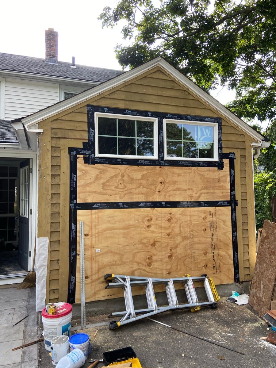 An exterior wall of a house under renovation with plywood covering a section below two new windows and black flashing tape.