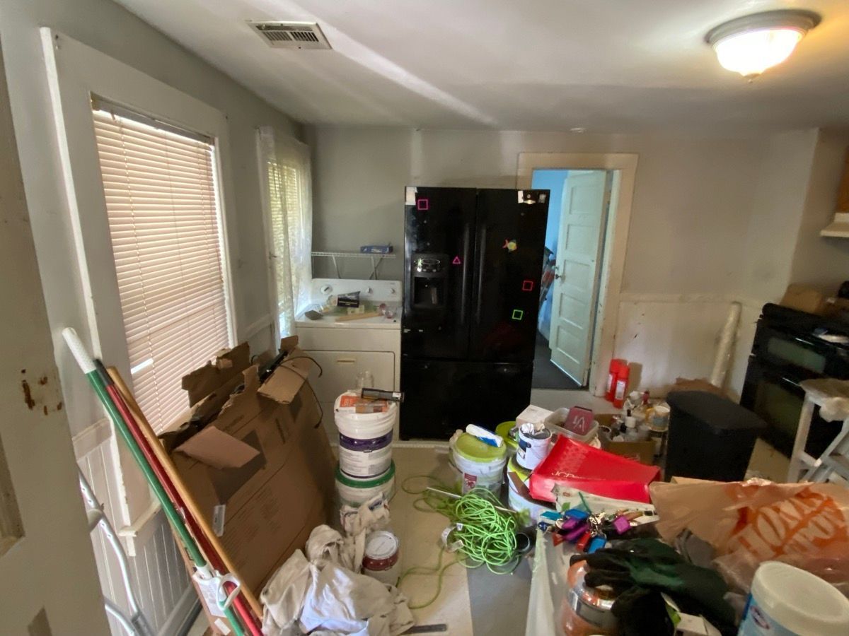 A cluttered kitchen undergoing renovation with a black refrigerator, cardboard boxes, and miscellaneous supplies.