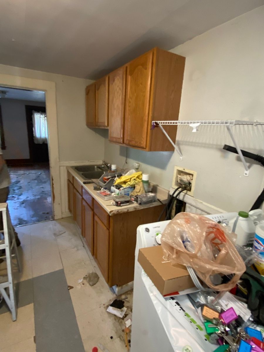 A kitchen under renovation featuring wooden cabinets, a sink, and a washer covered in clutter with bare flooring.