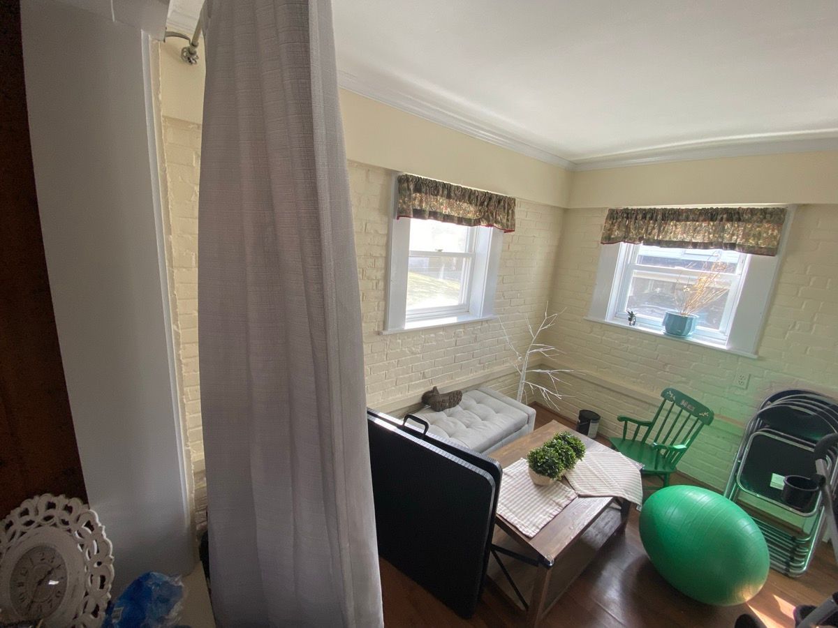 A room with beige walls, two windows with valances, a table with a plant, a green chair, a green exercise ball, and a cot.