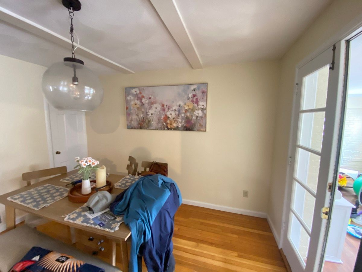 A light-filled dining area with a wooden table, a clear glass pendant light, and floral wall art on a cream-colored wall.