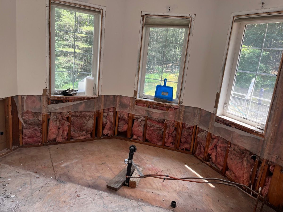 Renovated room showing wall studs, pink fiberglass insulation, three windows, and exposed plumbing on the floor.