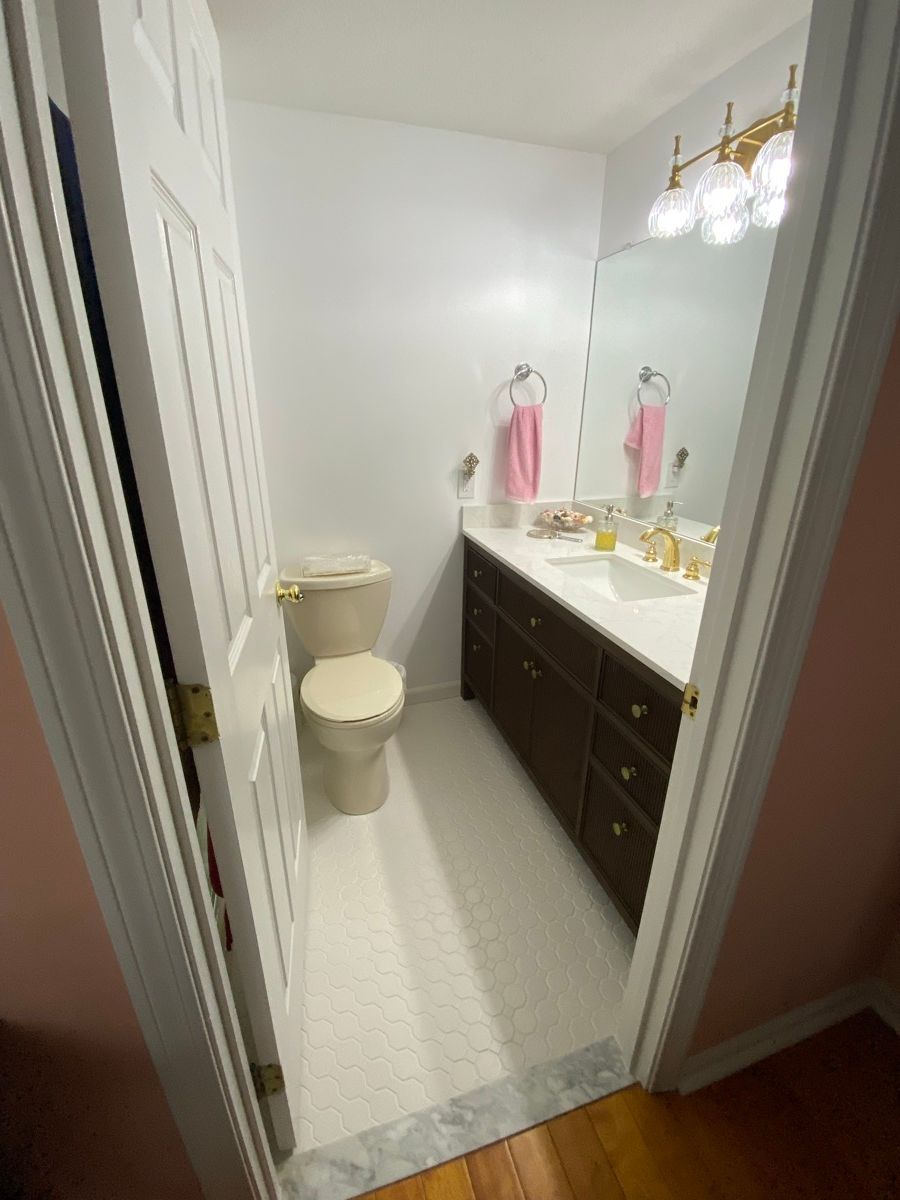 A small bathroom with white walls, a white toilet, a dark brown vanity with a white countertop, and a vanity light fixture.
