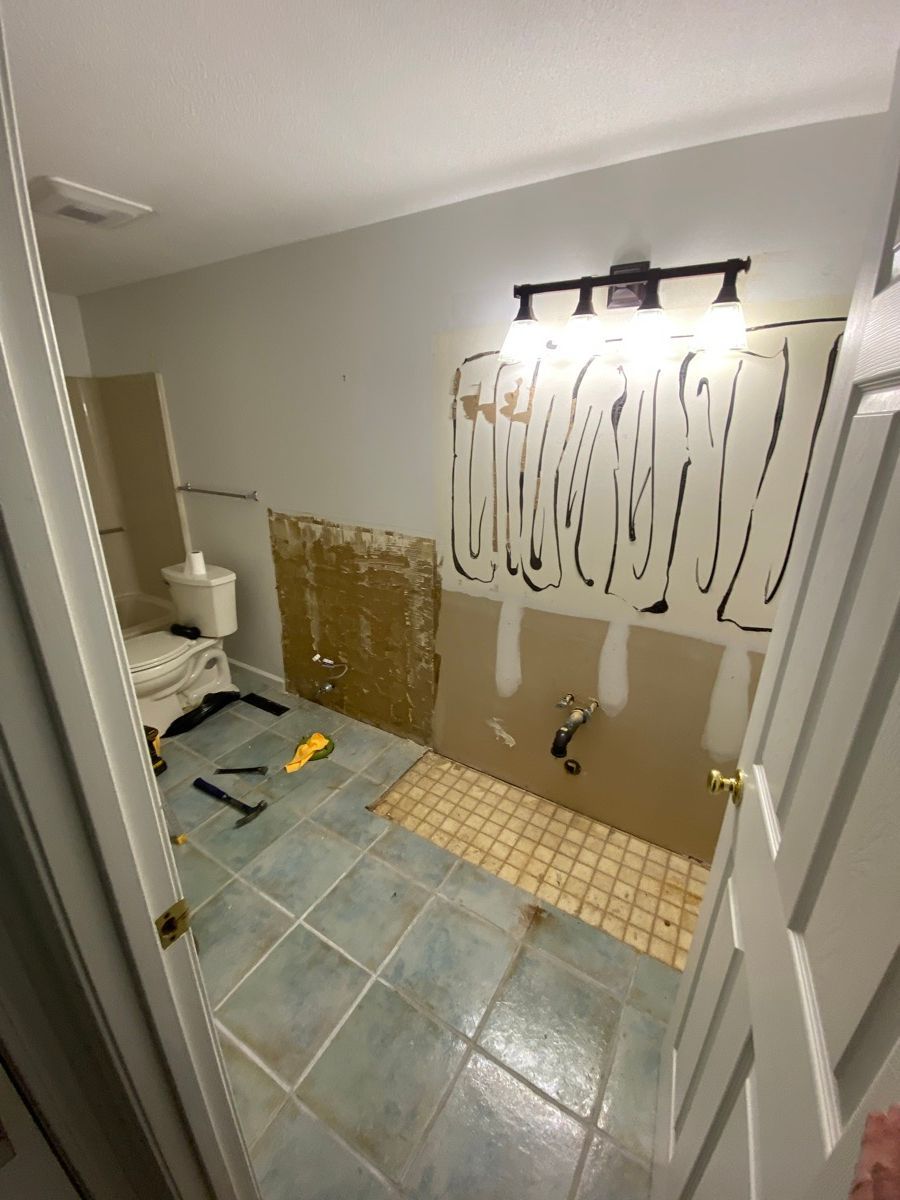 A bathroom under renovation with light blue tiled flooring, bare drywall with adhesive marks, and a wall-mounted light.