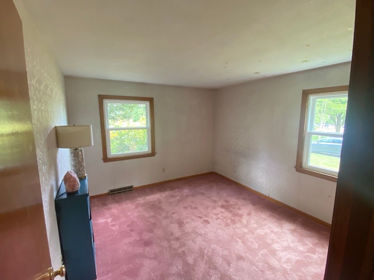 An empty bedroom with pink carpet, white walls, and two windows. A lamp sits on a blue cabinet in the corner.