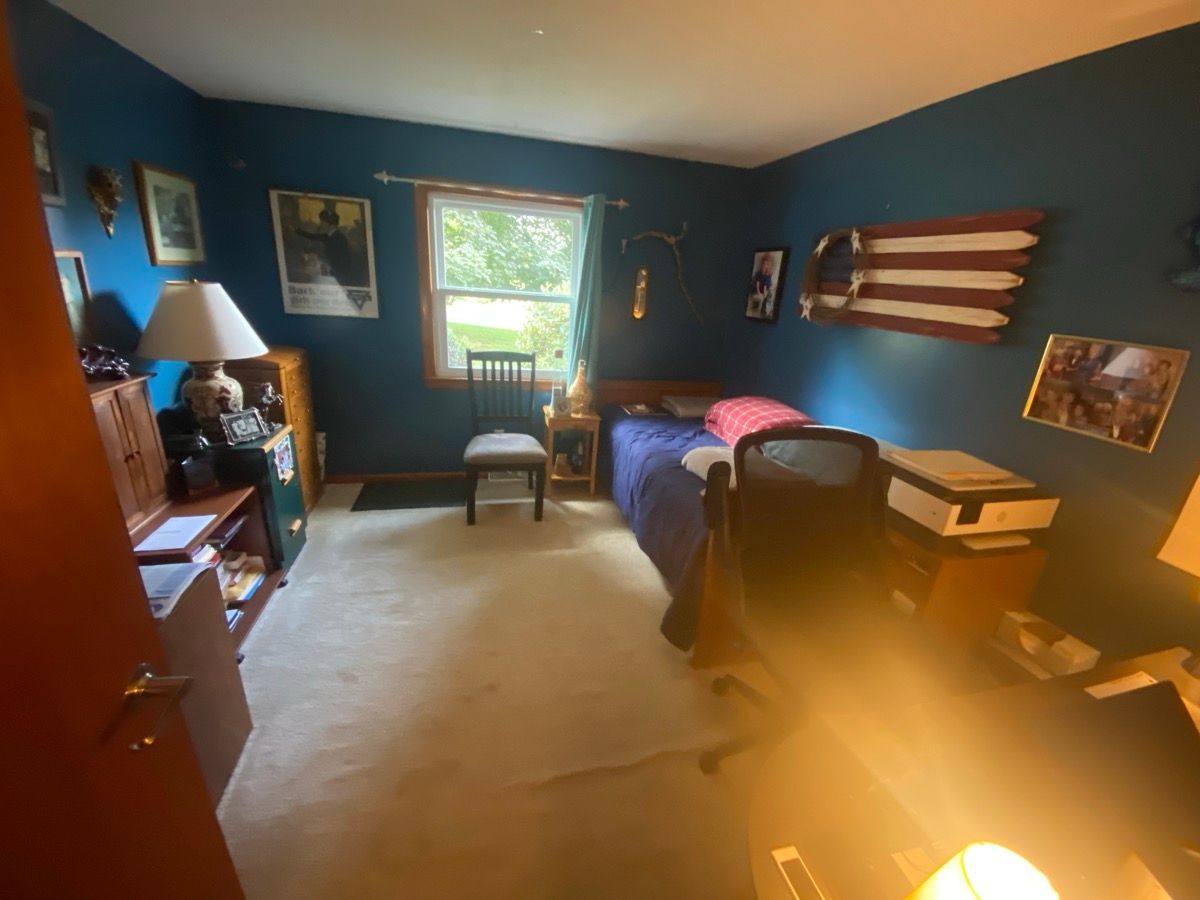 A blue-walled bedroom with a bed, a lamp on a desk, a window with a chair, and a wall-mounted wooden flag decoration.