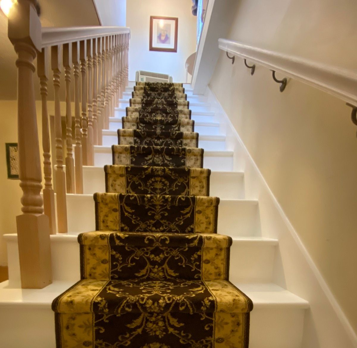 A staircase featuring white steps with a central brown and gold patterned runner, a wooden banister, and a wall handrail.
