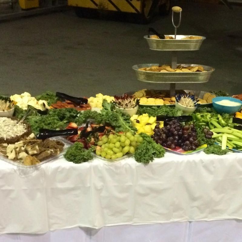 A catered buffet table with a three-tier server, fresh fruit, crackers, vegetables, and bowls of dips on a white cloth.