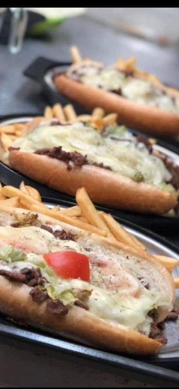 Three sandwiches filled with meat, melted cheese, and vegetables, served with a side of french fries on black trays.