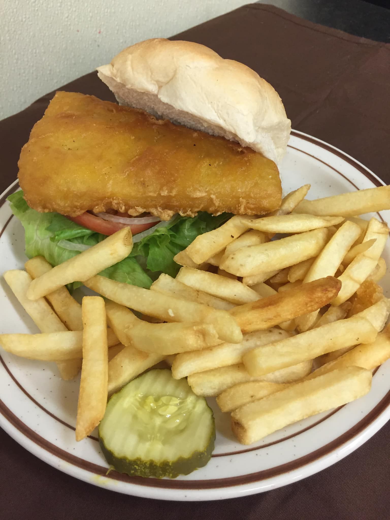 A breaded fish sandwich on a bun with lettuce and tomato, served with French fries and a pickle slice on a plate.