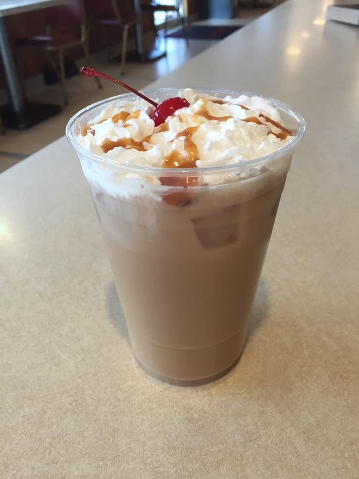 A clear plastic cup of iced coffee topped with whipped cream, caramel drizzle, and a maraschino cherry.