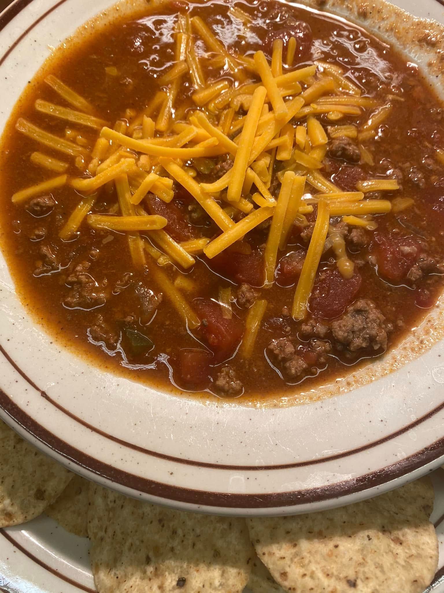 A bowl of red chili topped with shredded cheddar cheese, served with round tortilla chips on a white plate.