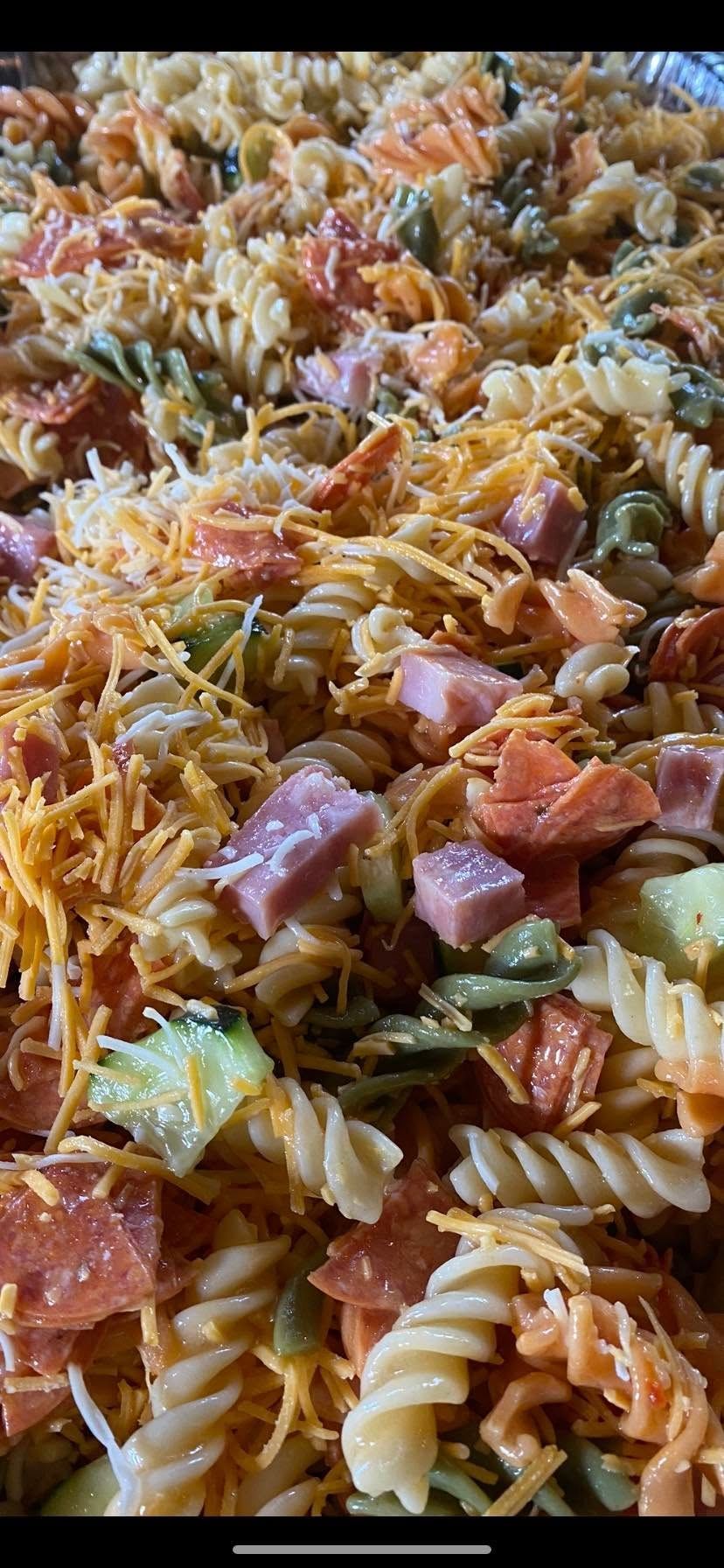 A close-up view of pasta salad with rotini, cubed ham, shredded cheddar cheese, tomato pieces, and chopped cucumber.