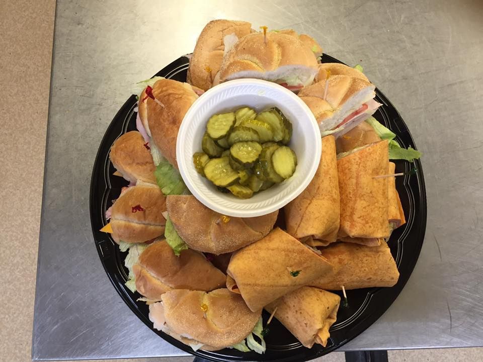A black plastic platter holding a bowl of pickle slices surrounded by an assortment of sandwich halves on a metal table.