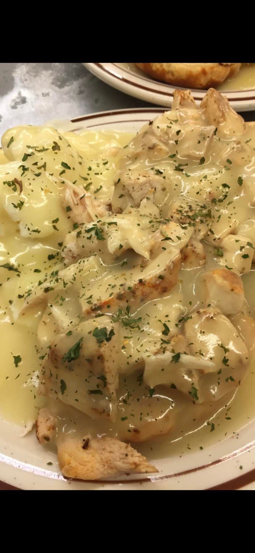 A close-up of a chicken dish topped with a creamy sauce and herbs, served with potatoes on a white plate with a brown rim.