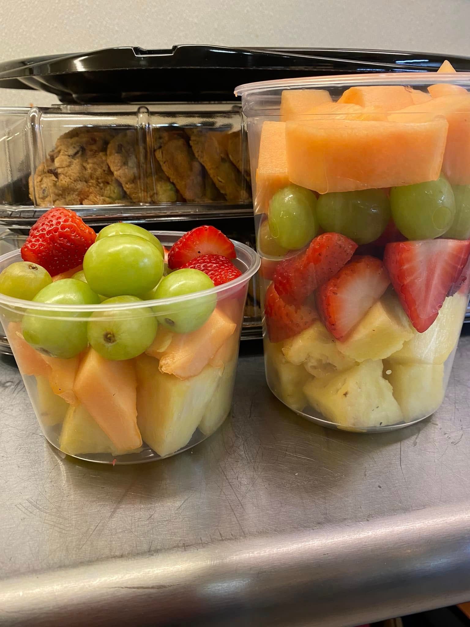 Two plastic cups filled with cut pineapple, strawberries, melon, and green grapes, with cookies in the background.