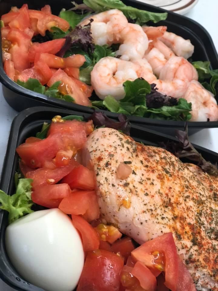 Two black take-out containers each holding a salad with fresh tomatoes, greens, and either shrimp or seasoned chicken.