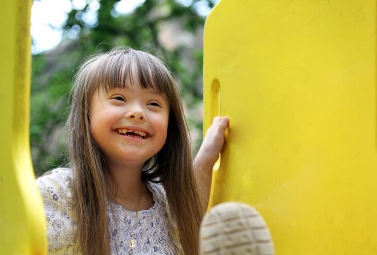 Smiling child with long hair peeks from behind a bright yellow playground structure outdoors