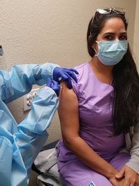Vaccinated woman in purple scrubs receiving a shoulder injection from a gloved healthcare worker