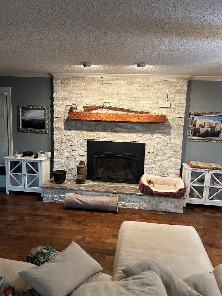 Cozy living room with stone fireplace, mounted TV, rustic mantel, and cushioned chairs around a hardwood floor.