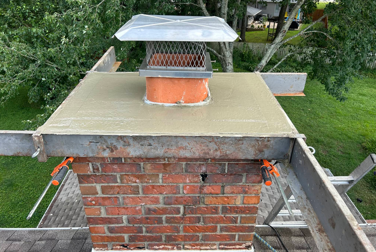 Brick chimney with metal cap on a roof, surrounded by green yard and scaffolding.