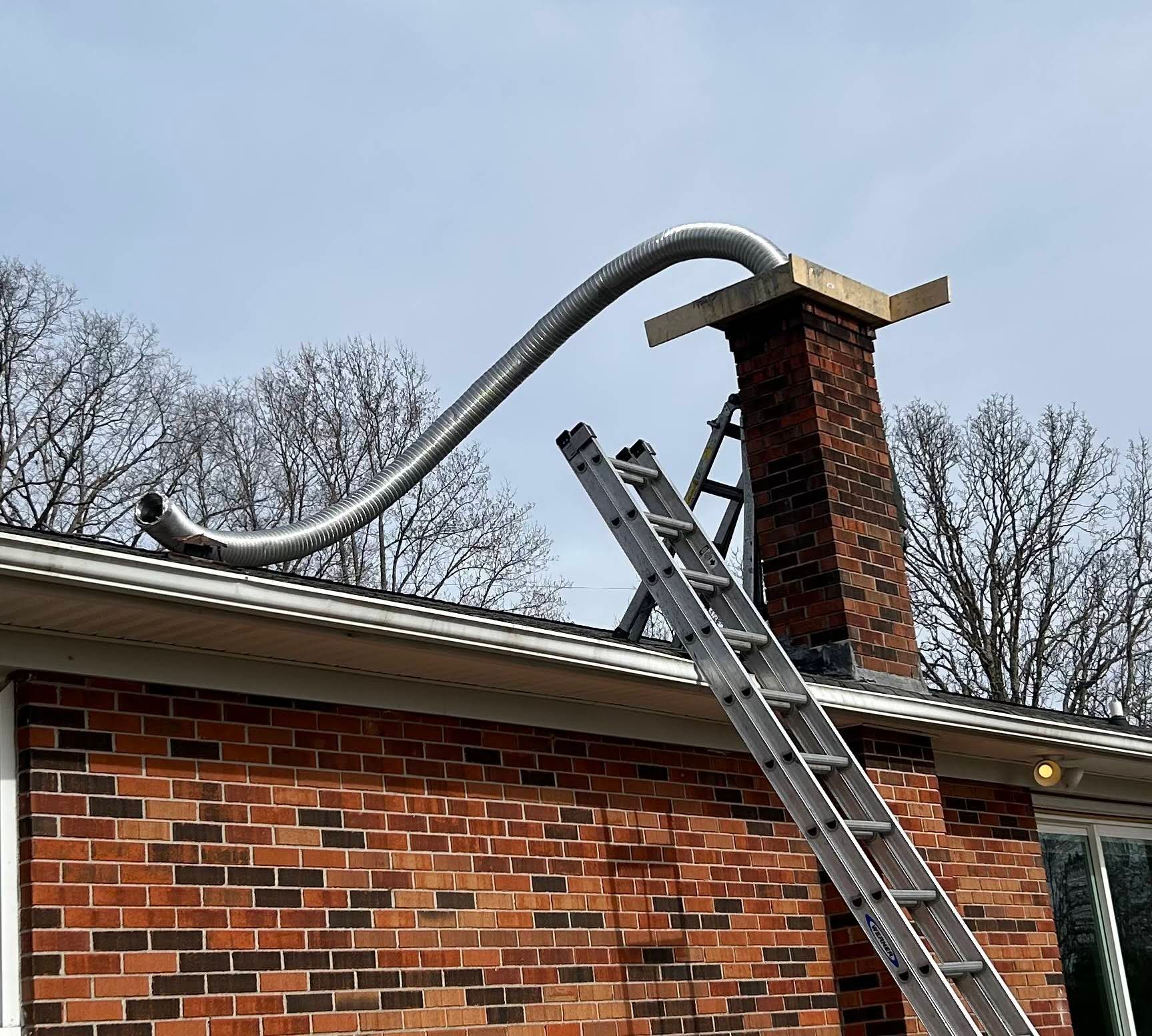Roofing crew installing a flexible chimney vent through a brick chimney, with a ladder on the roof