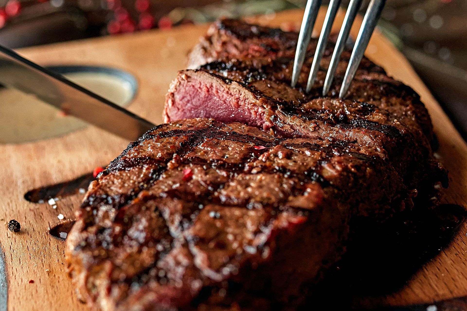 Sliced grilled steak on a wooden board, being pierced by a fork. A small dish of sauce is visible.
