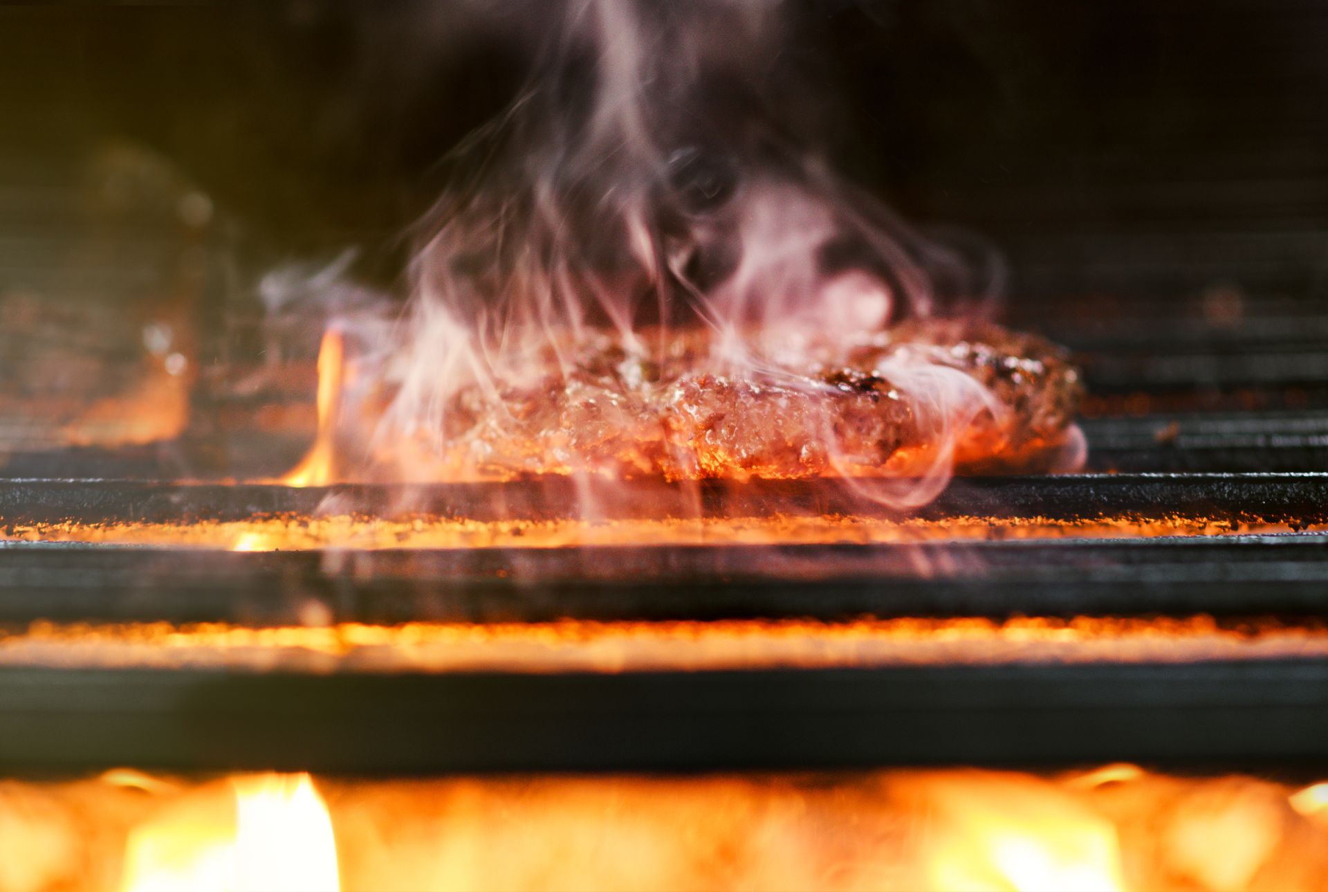 Hamburger patty sizzling on a hot grill, flames visible, smoke rising.