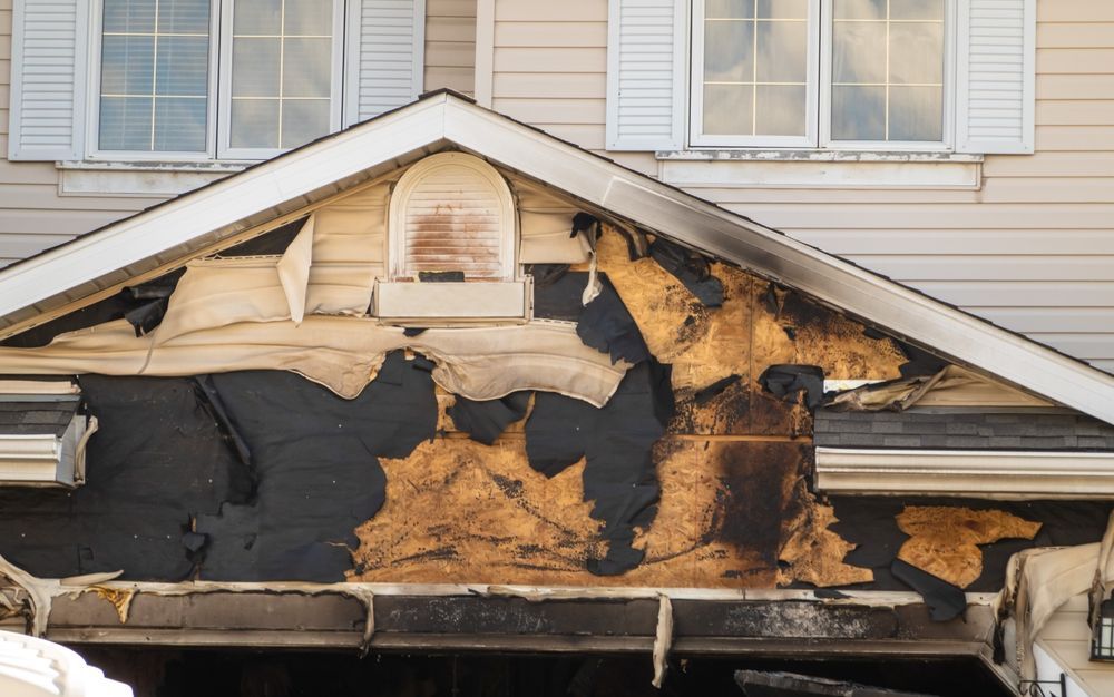 Severely fire-damaged house exterior with charred siding and exposed insulation beneath the roofline