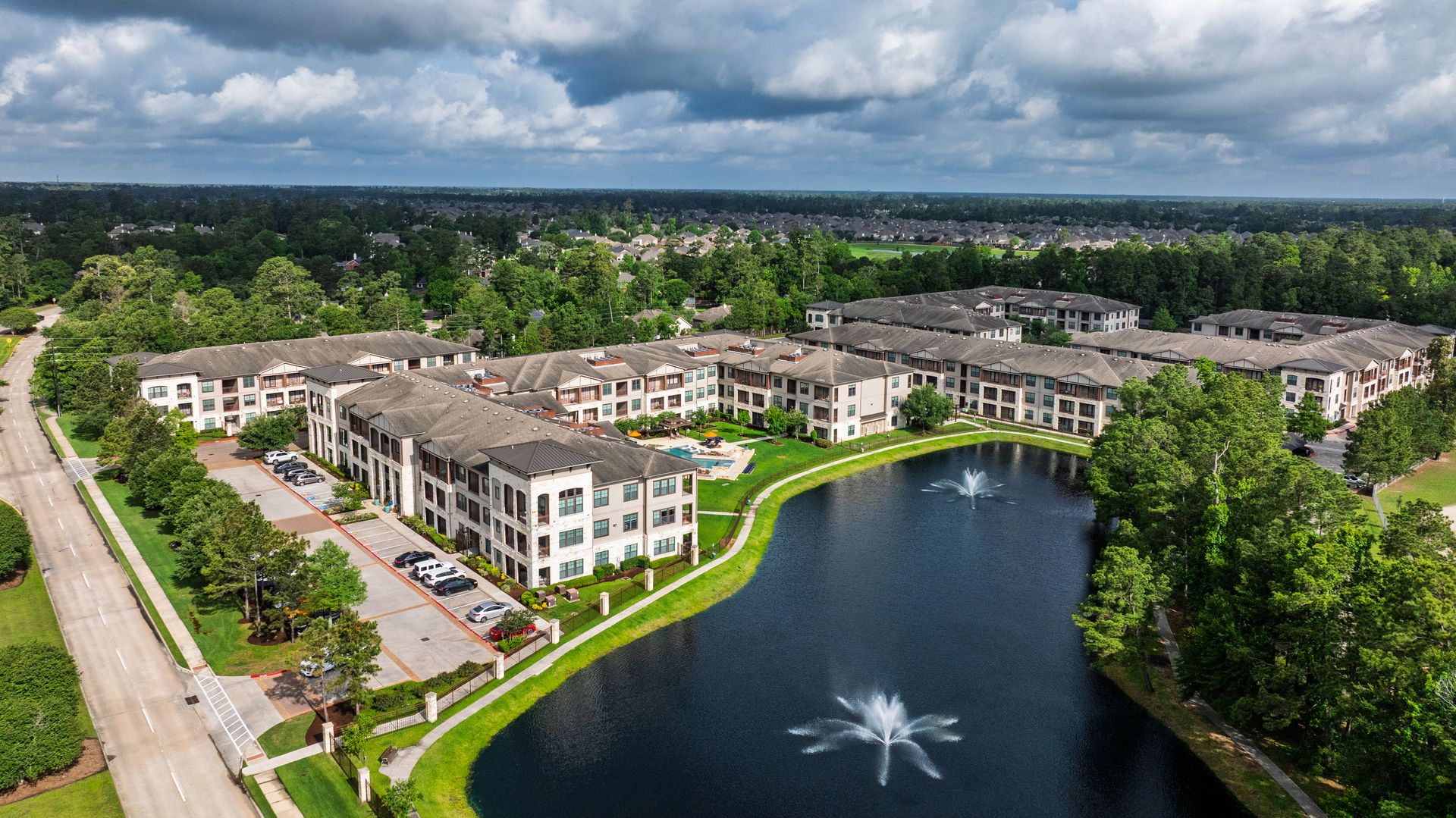 Aerial view of apartment buildings around a pond with a fountain, trees, and a nearby road at Olympus Auburn Lakes in Spring, TX.