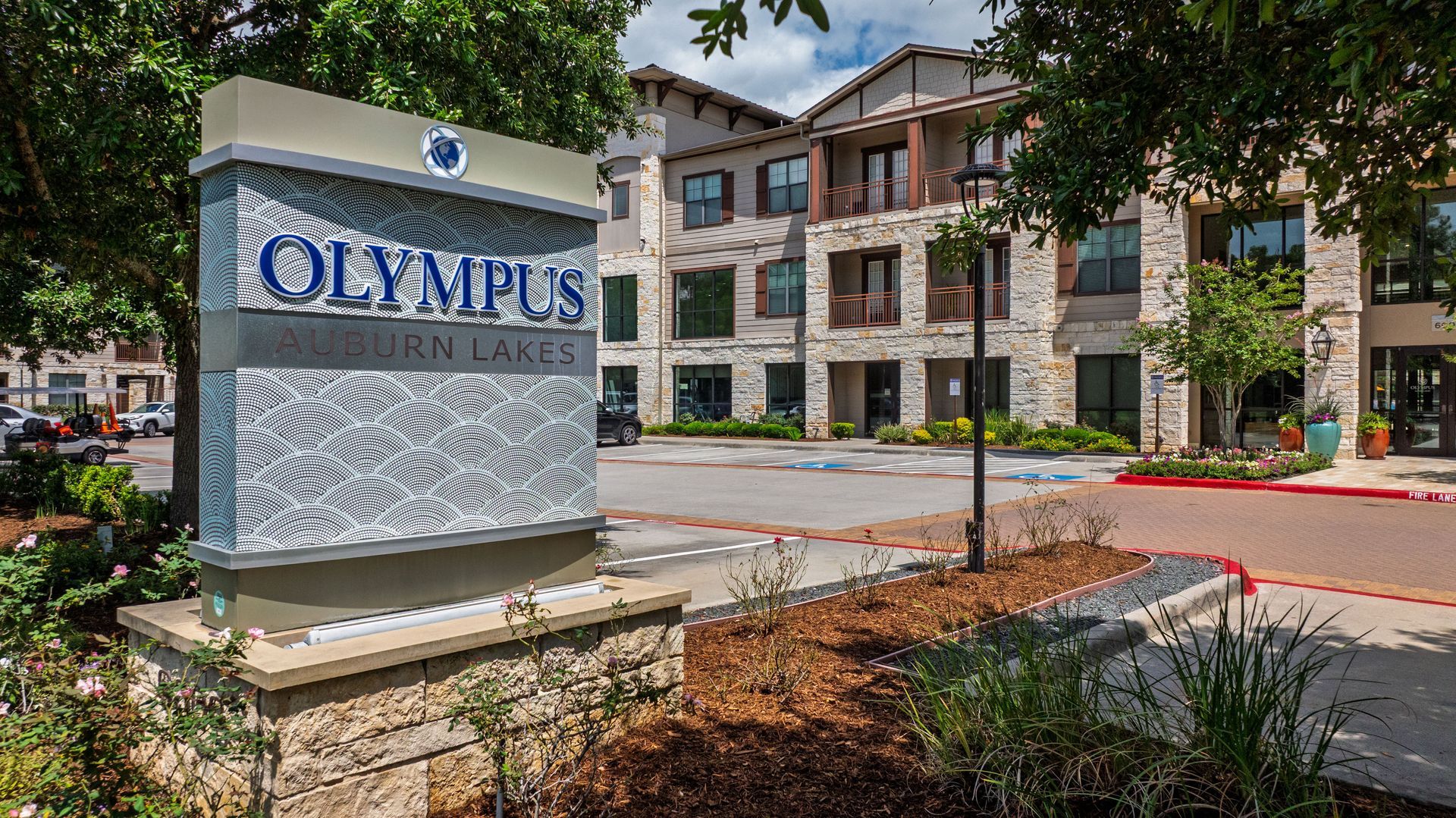 Stone sign for “Olympus at Enclave” outside a modern apartment building with trees and landscaping at Olympus Auburn Lakes in Spring, TX.