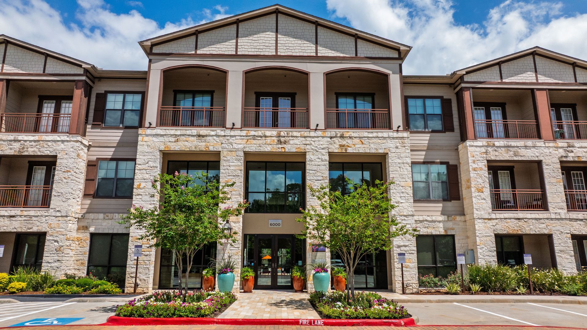 Modern stone apartment building entrance with balconies, glass doors, and flower beds at Olympus Auburn Lakes in Spring, TX.