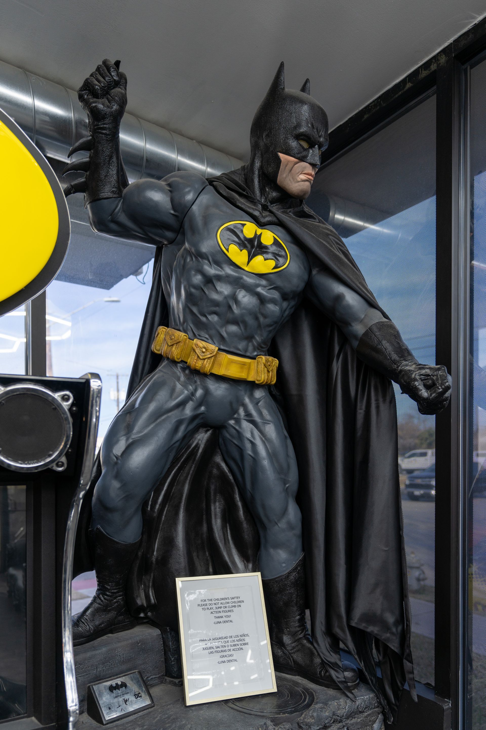 Batman statue standing indoors with raised arm, black suit, yellow emblem, and cape