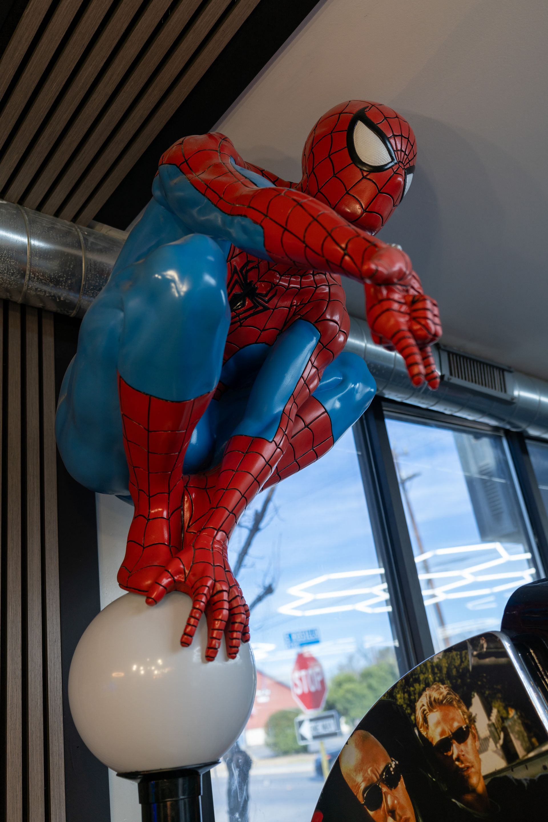 Spider-Man balloon figure posed crouching indoors near a window, with red and blue suit and a white base