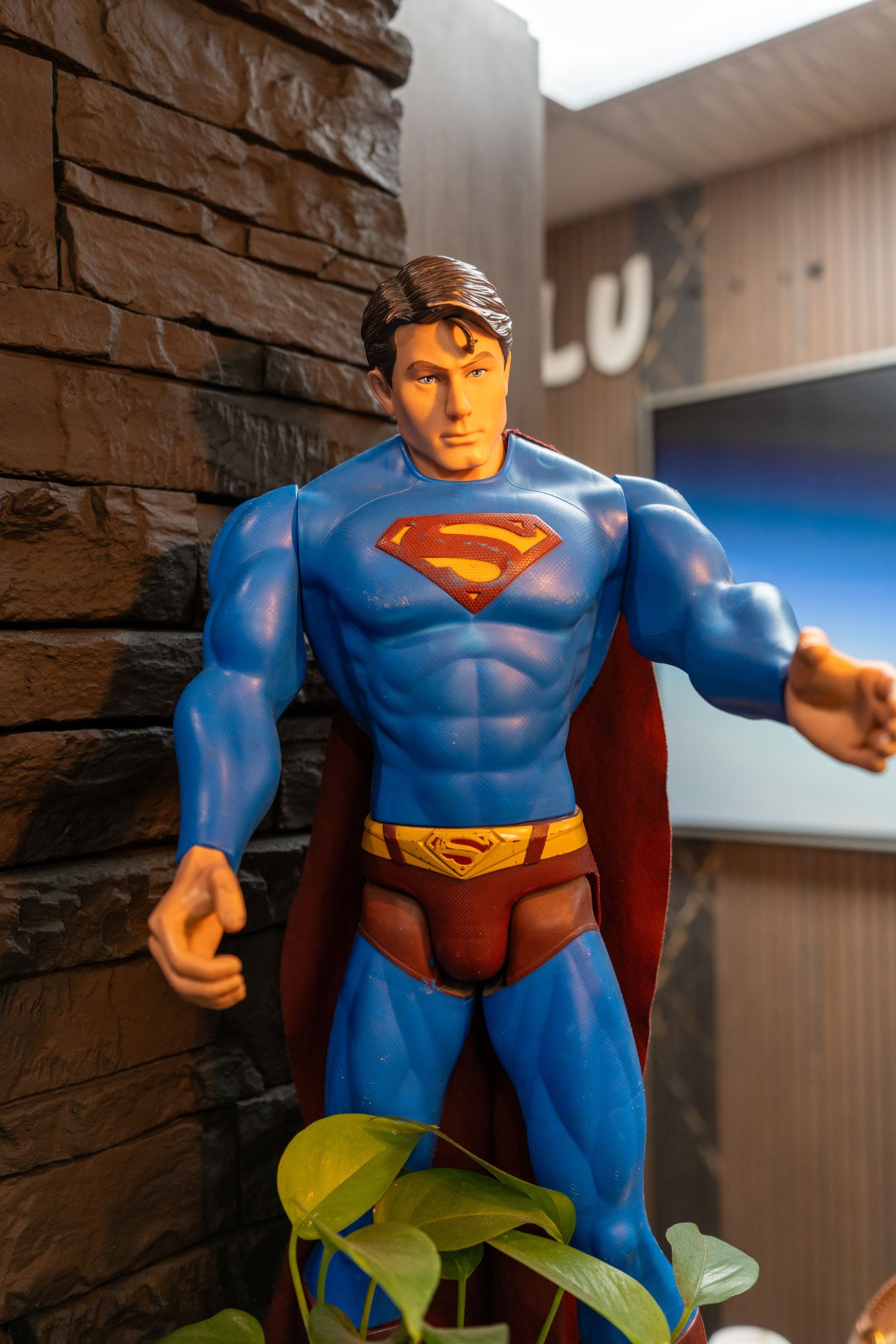 Superman action figure in a blue suit standing beside a brick wall with a plant in the foreground