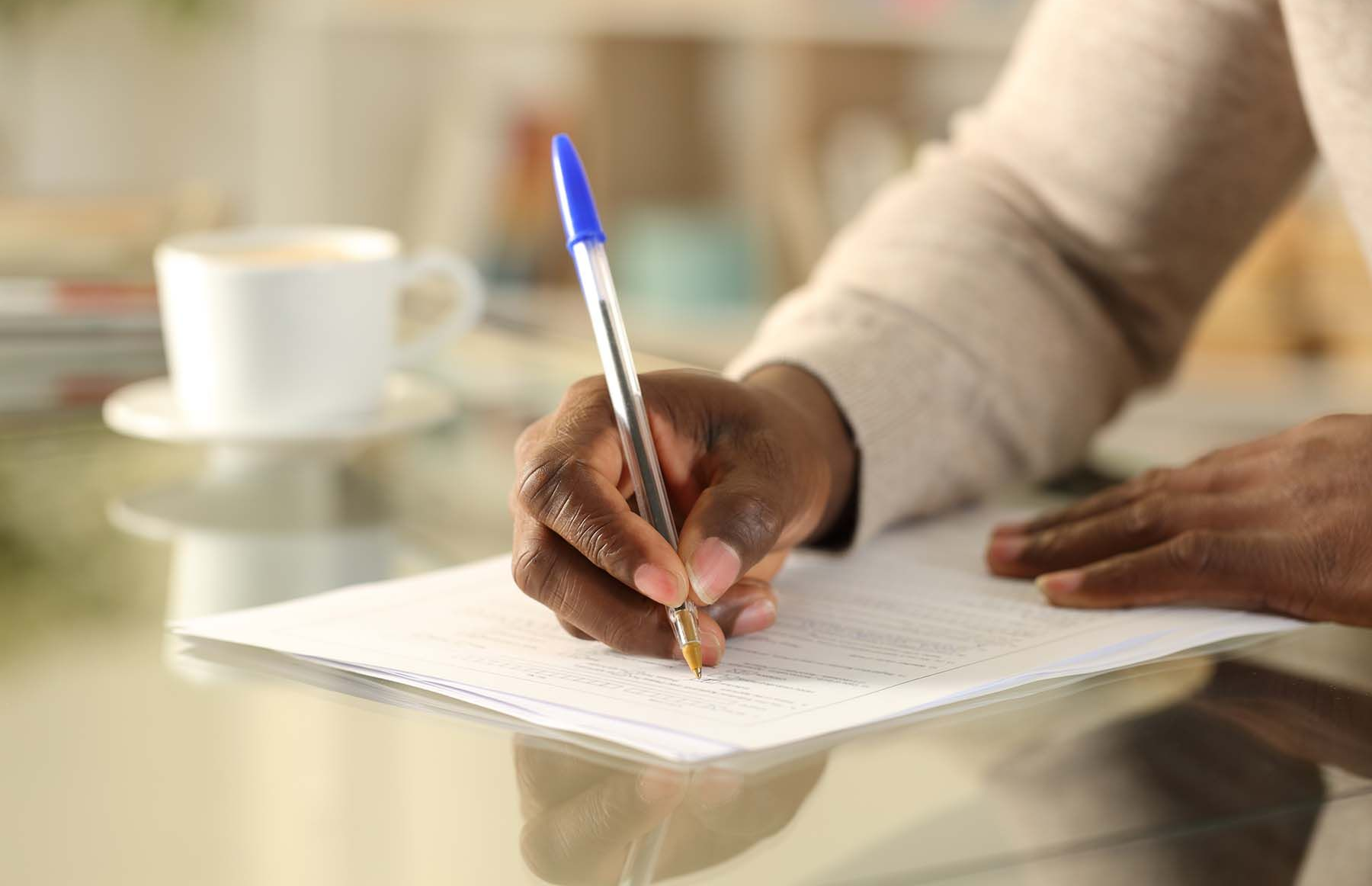 Person's hand writing with blue pen on white paper at a table. A coffee cup is in the background.