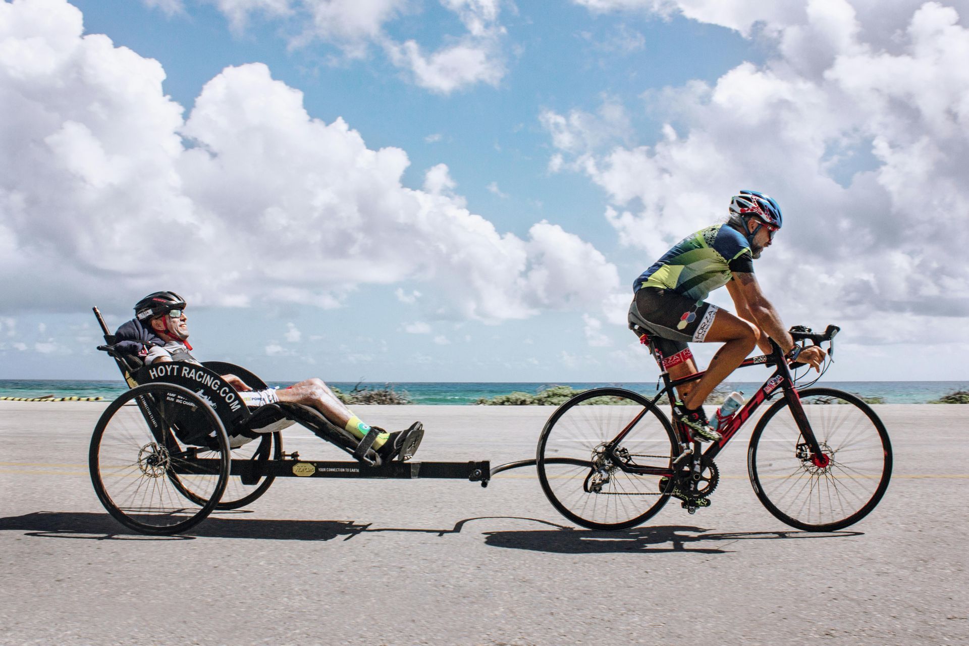 Two cyclists on a beach road, one riding a recumbent bike under a bright, cloudy sky.