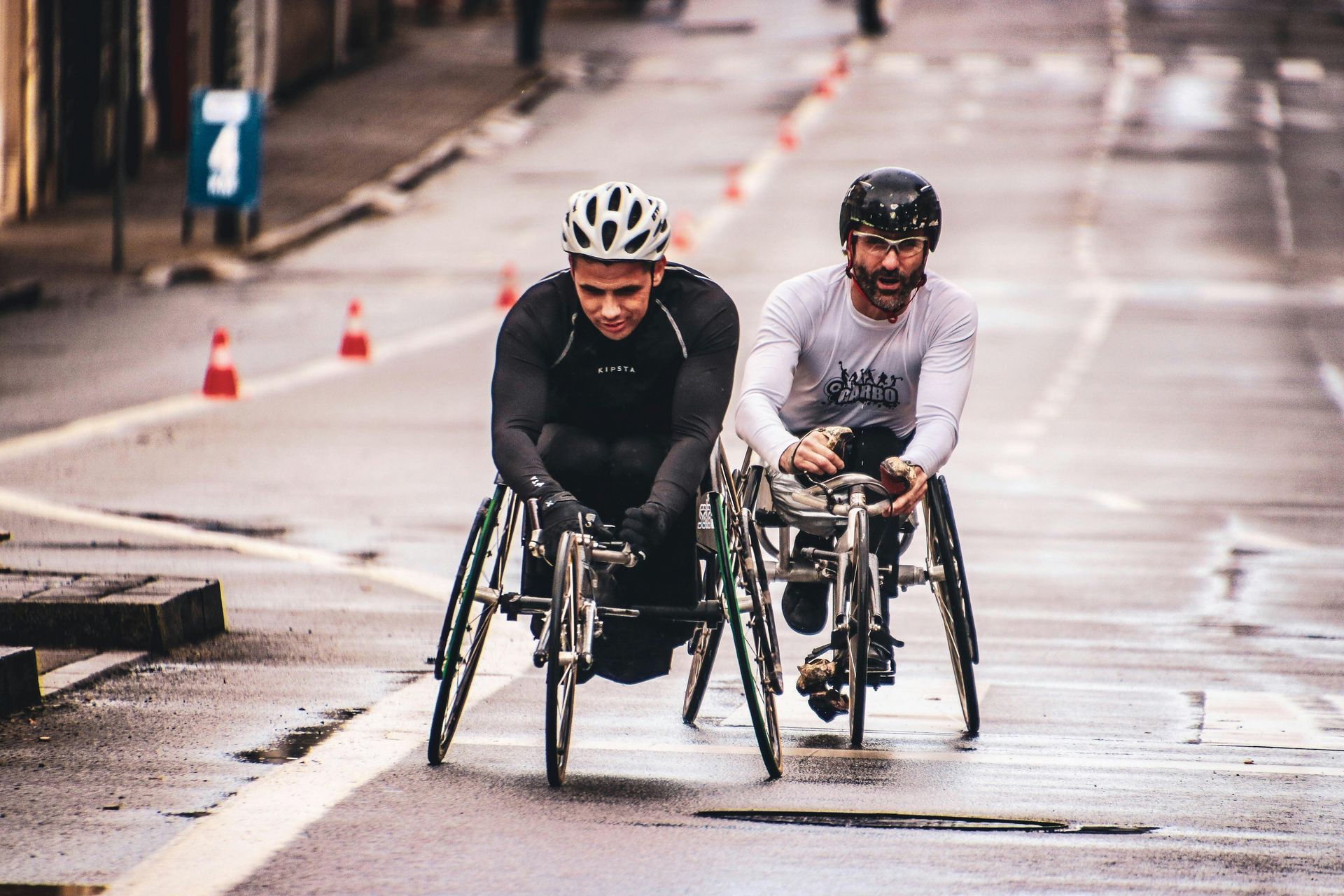 Two cyclists in helmets race side by side on a wet street with cones in the background