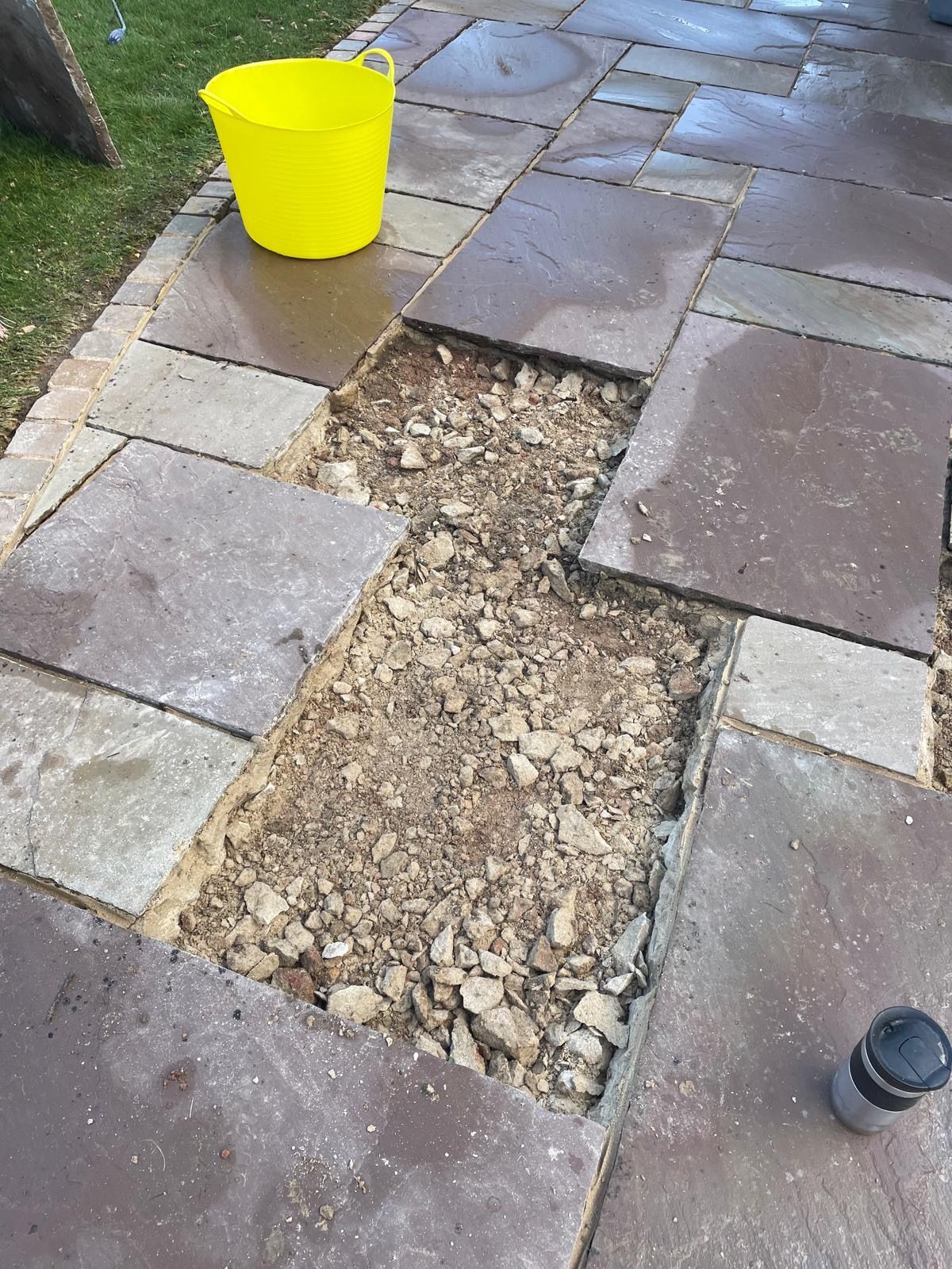 A patio area with two missing paving stones, revealing a gravel base, next to a yellow bucket and a small black container.