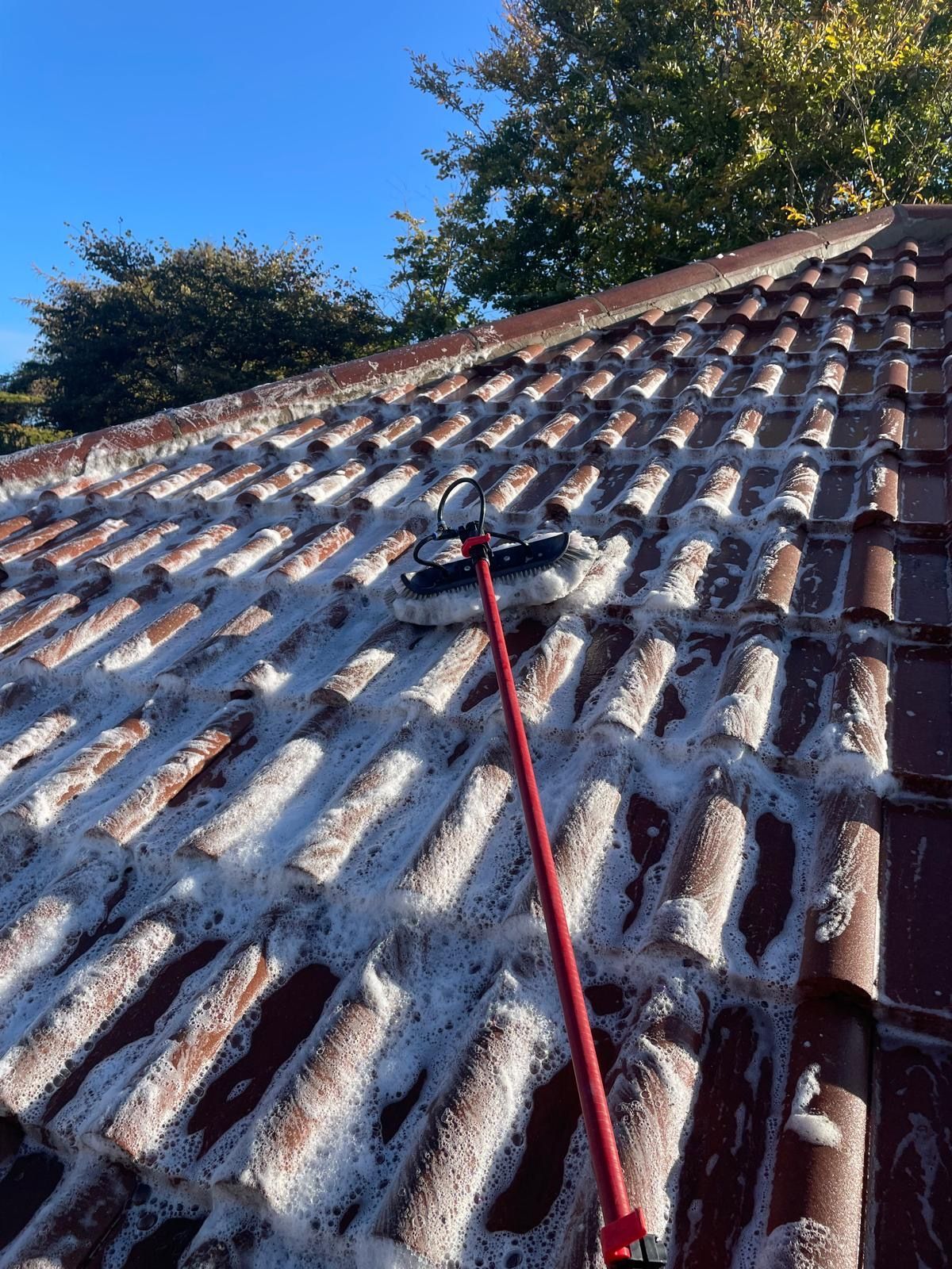 Roof being cleaned with a long-handled brush; red tiles covered in white foam under a blue sky.