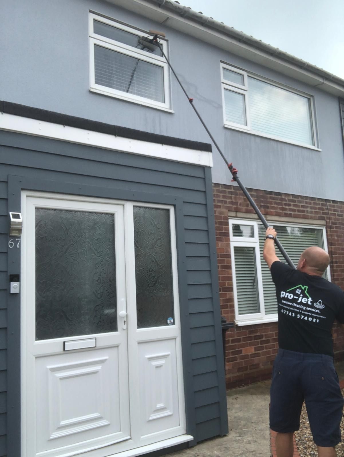 Man cleaning a two-story house's windows with a long-handled brush. Gray siding, brick, and white door.