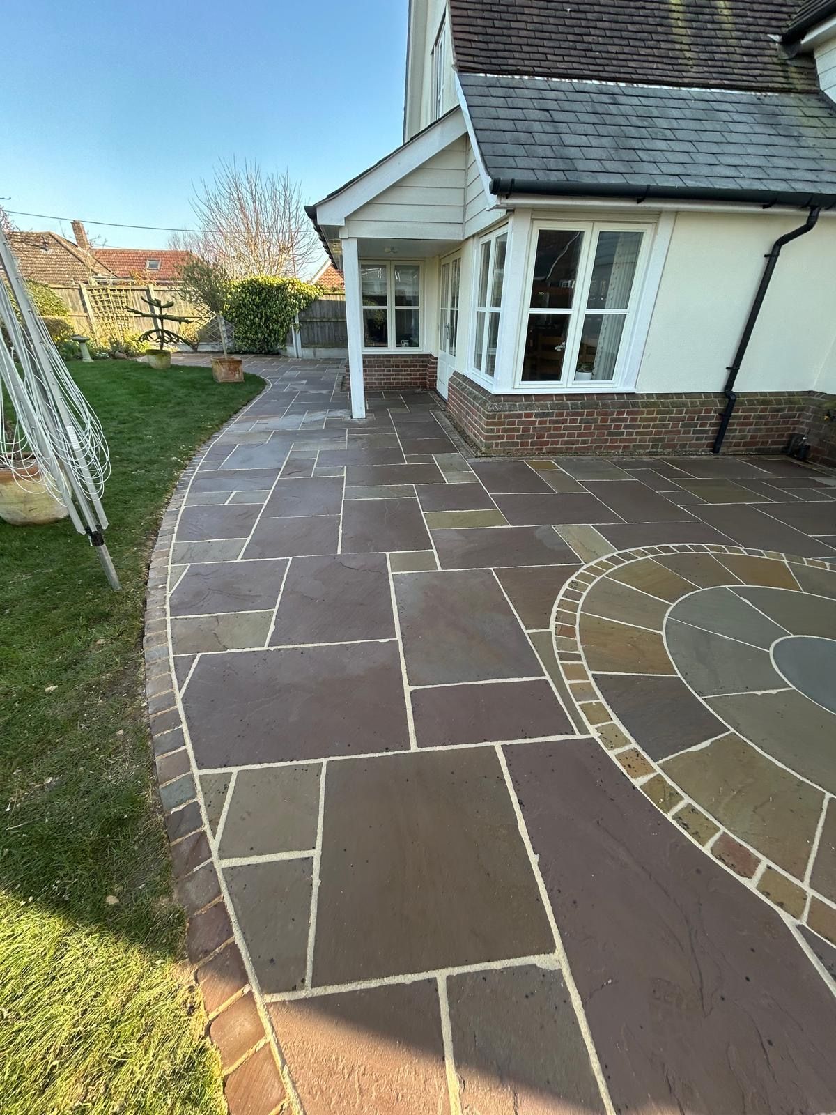 A newly laid patio with brown stone slabs in a circular design next to a white house with a dark roof and brick foundation.