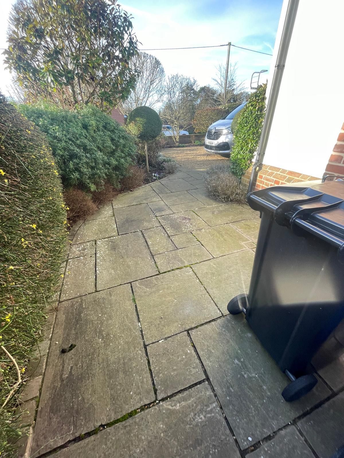A stone-paved driveway leads past a large hedge to a parked car on a sunny day, with a trash bin in the foreground.