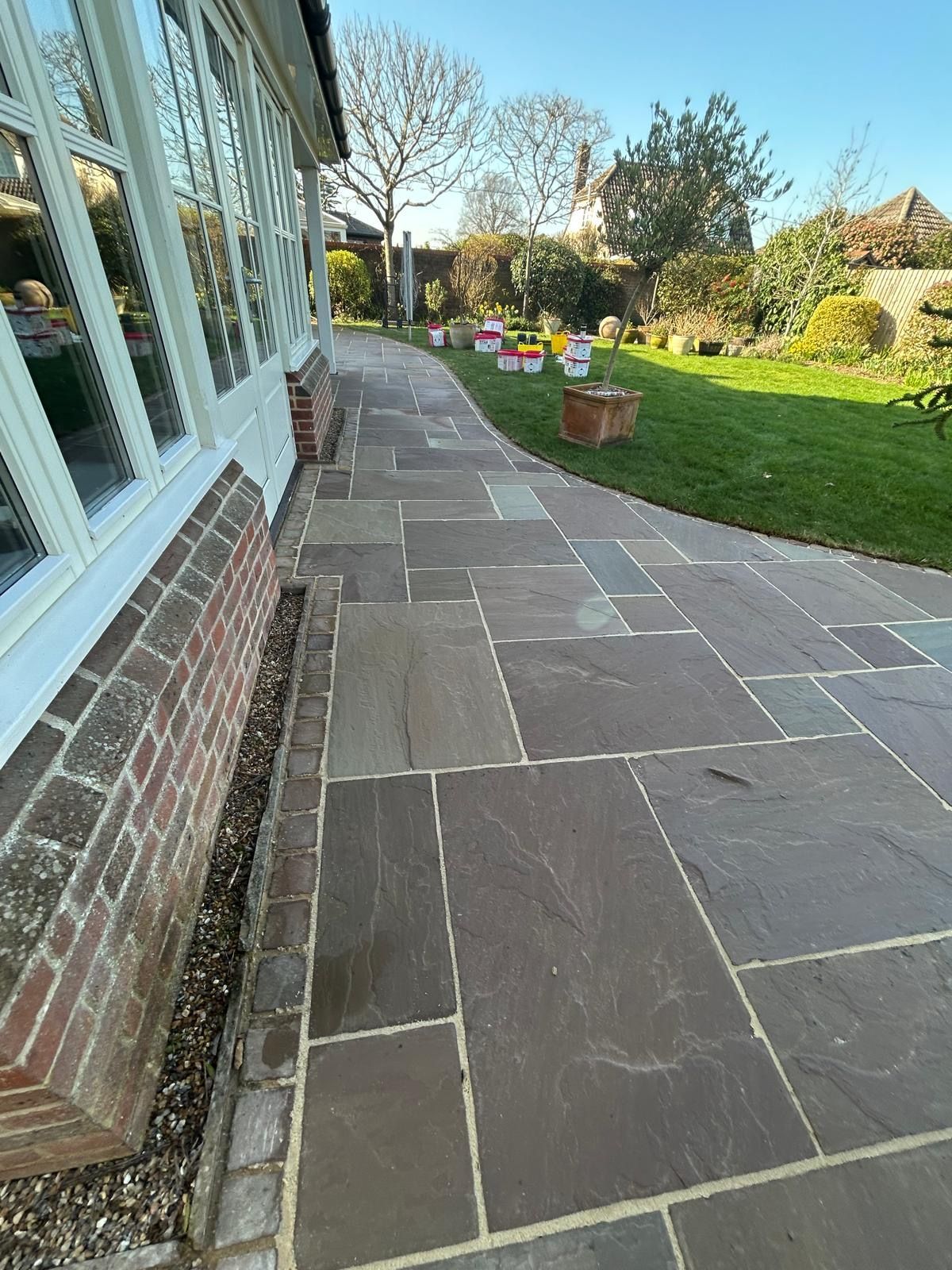 A stone patio path runs alongside a brick-walled sunroom, leading toward a grassy backyard on a sunny day.