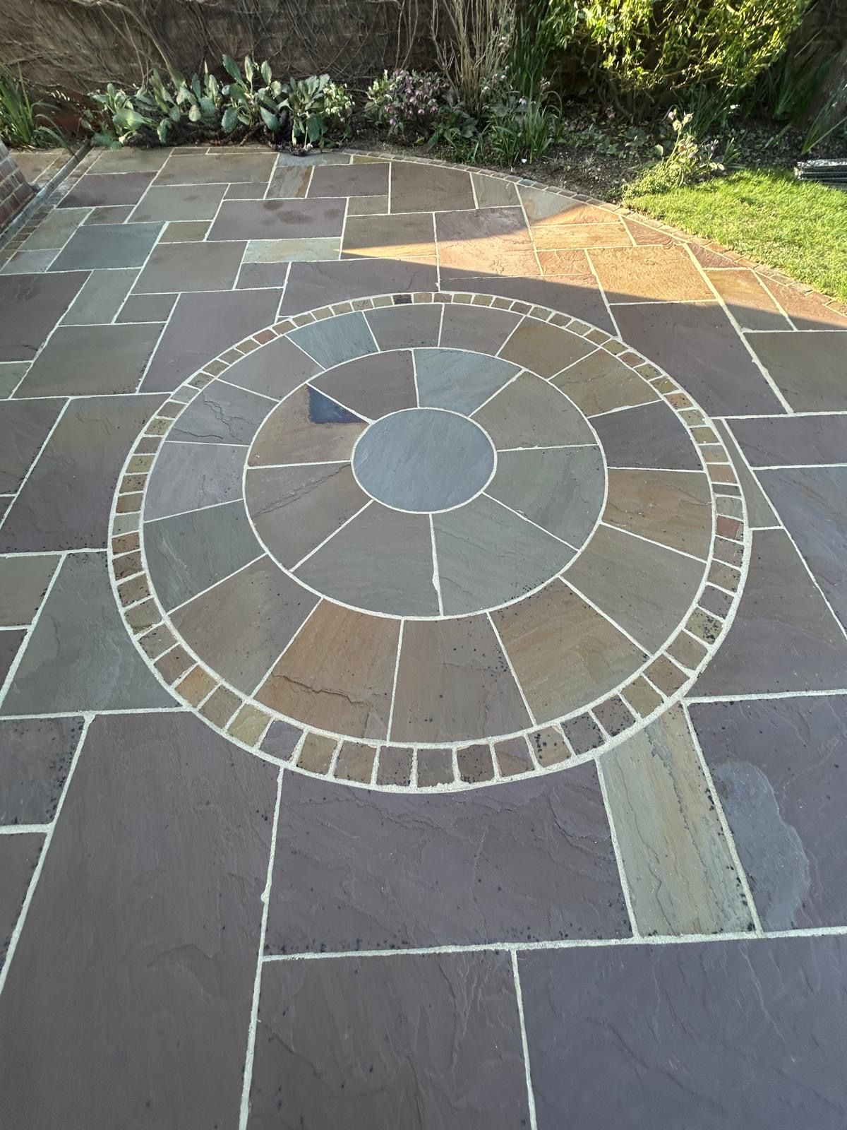 A circular stone patio feature with a grey center, surrounded by brown and tan radial pavers, set in a paved patio.