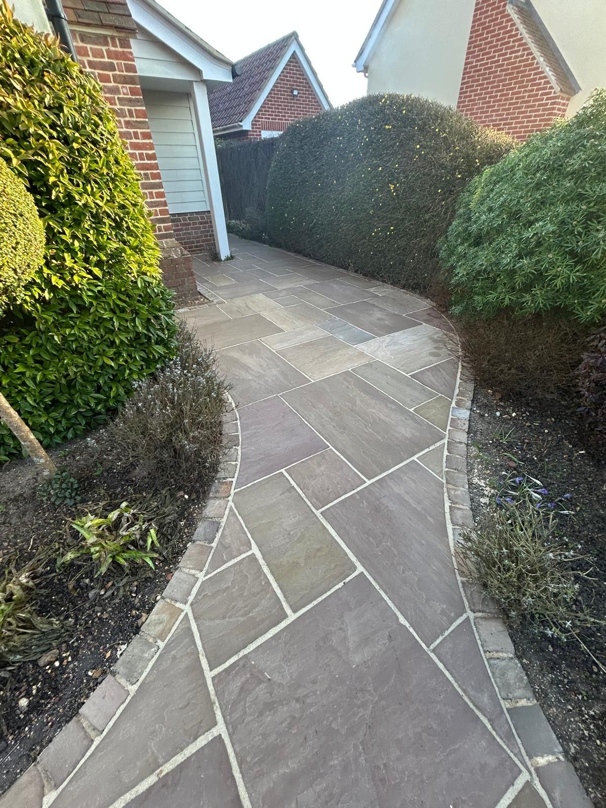 A stone garden path with dark borders winds between neatly trimmed hedges towards a house entrance.
