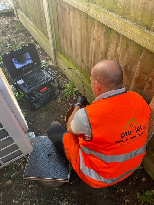 Man in orange vest inspects sewer with camera near a wooden fence. Camera displays pipe view.